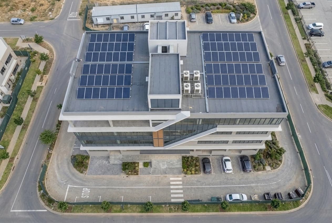 Straight-down aerial view of a completed commercial building: two roof-mounted solar arrays either side of a central plant enclosure, a bank of air-conditioning condensers, and the parking bays, drive, pedestrian crossing and landscaped forecourt laid out around the building.