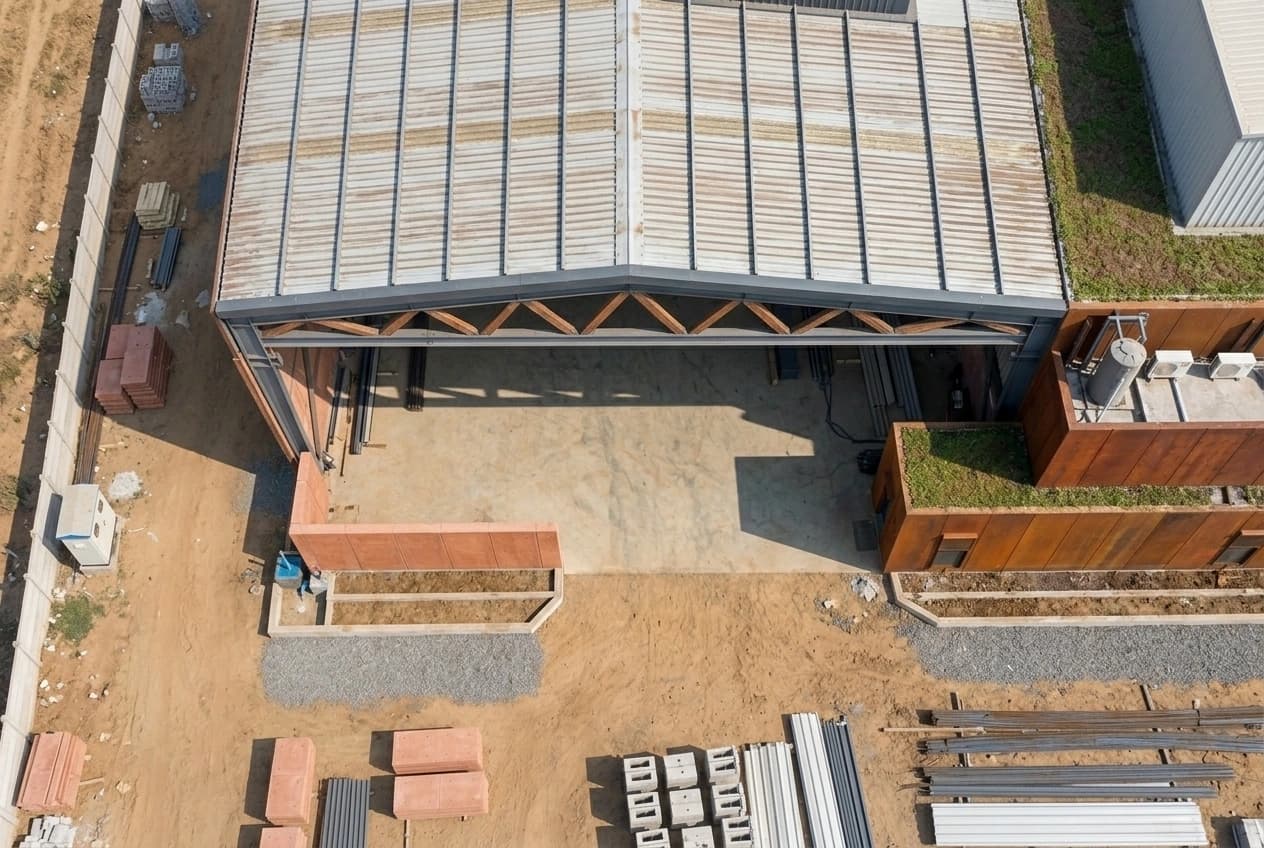 Straight-down aerial view of an industrial unit under construction, showing the roof sheeting and ridge, the open structural bay below, the concrete hardstanding at the entrance and building materials laid out on the surrounding ground.